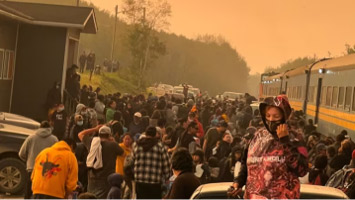 A picture of a crowd of people waiting to board a KRC train to escape from wildfires threatening their community. The sky is hazy and orange from smoke.