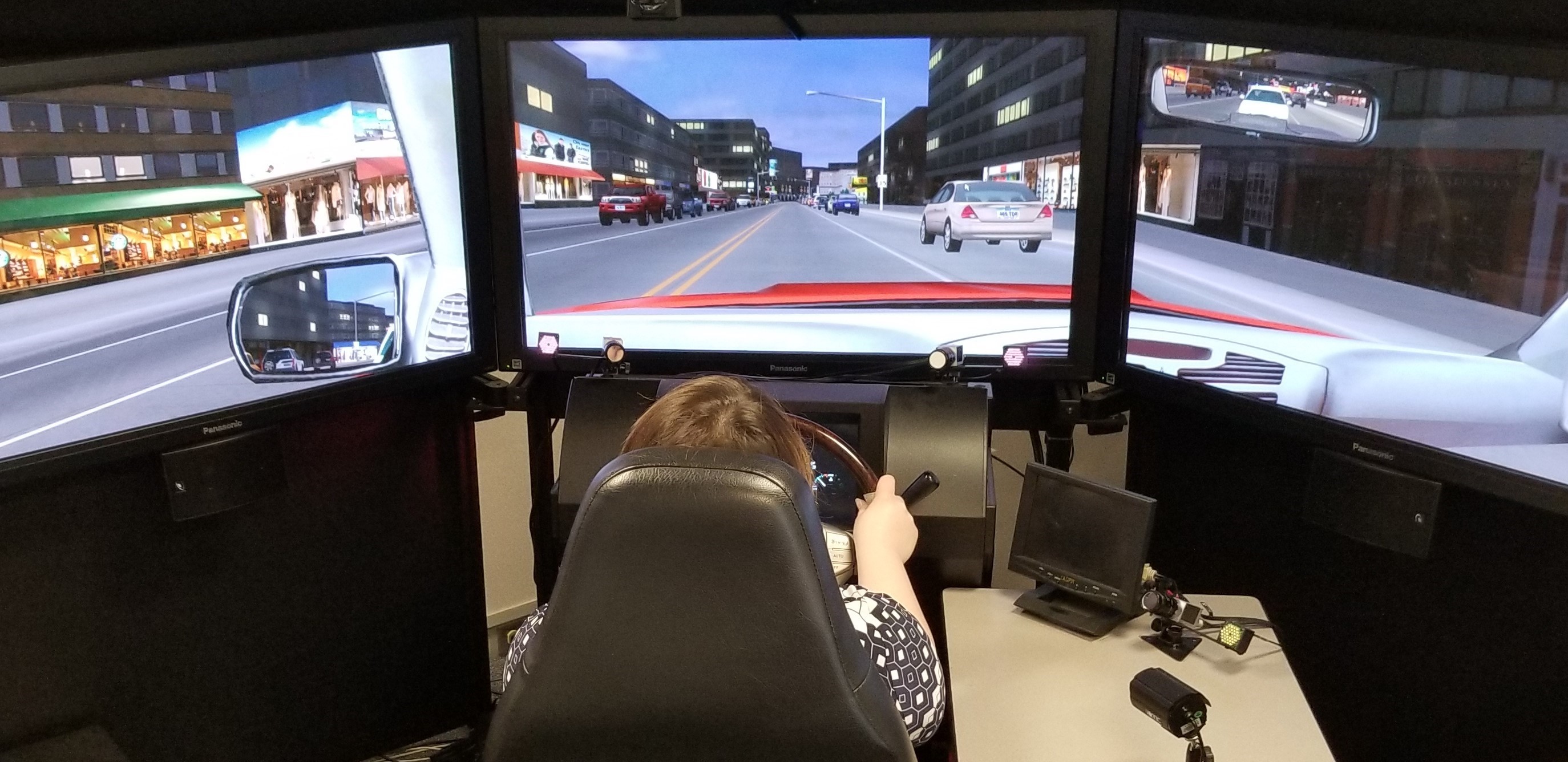 Woman using a driving simulator. Three screens display a panoramic view of a city road. Her hands are on the wheel.